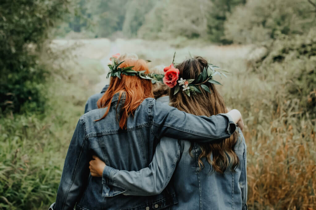 two women wearing denim jackets with flowers in their hair walking in a meadow with their arms around each other