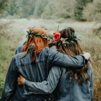 two women wearing denim jackets with flowers in their hair walking in a meadow with their arms around each other